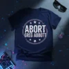 Navy blue t-shirt with "ABORT GREG ABBOTT" text in a distressed, glitchy font within a circular graphic with stars