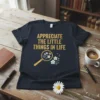T-shirt with gold text "APPRECIATE THE LITTLE THINGS IN LIFE" above a magnifying glass focusing on small flowers and a