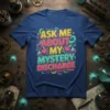 Blue t-shirt with bold, colorful text: "ASK ME ABOUT MY MYSTERY DISCHARGE" in a playful font with science-themed graphics