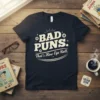 Dark t-shirt with white text reading "BAD PUNS. That's How Eye Roll." featuring eye graphics and playful typography