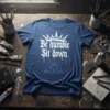 White "Be Humble Sit Down" text and crown graphic on a blue t-shirt, with two figures meditating below