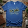 Blue t-shirt with "be you. The world will adjust." in gothic and simple fonts, framed by abstract golden geometric lines