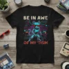 Robot cat wearing sunglasses with text "BE IN AWE OF MY 'TISM" on a black t-shirt, set against a circuit