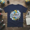 BE THE CHANGE quote in bold white lettering with a yellow and blue abstract phoenix graphic on a navy t-shirt