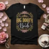 Black t-shirt with "BIG BOOTY Bride" text in gold glitter and white script, surrounded by decorative floral elements and a