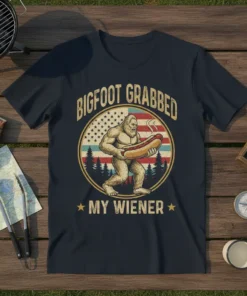 Bigfoot holding a hot dog with the text "BIGFOOT GRABBED MY WIENER" above an American flag background and pine trees