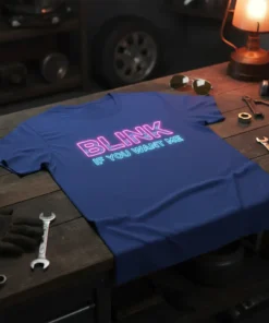 Blink If You Want Me quote in neon pink and blue on a navy blue t-shirt, styled on a workbench