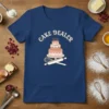 Navy blue t-shirt with "CAKE DEALER" text above a tiered cake graphic, rolling pin, and whisk