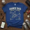 Blue t-shirt with yellow text "DANCE DAD COMP DAY PLAYBOOK" and a dance competition playbook diagram with terms like "Build
