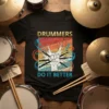 Black t-shirt with "Drummers Do It Better" text and a graphic of a fist holding drumsticks, surrounded by colorful abstract