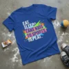 Blue t-shirt with white text "EAT. SLEEP. TAKE KIDS TO SPORTS. REPEAT." and colorful paint splatters, including soccer balls