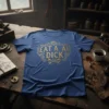 Blue t-shirt with gold "Eat A AI Dick" text and wrench graphic, framed by ornate filigree