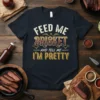 Black t-shirt with "Feed Me Brisket And Tell Me I'm Pretty" text in vintage distressed font, celebrating BBQ and brisket