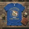 Blue t-shirt with gold text "FISH AROUND and FIND TROUT" and a graphic of a rainbow trout with fishing gear