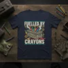 Fuelled By Crayons text design with a wooden crate filled with colorful crayons and small tools, printed on a navy
