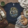 Navy t-shirt with "Gin Cats & Naps" text design in gold, cream, and blue, surrounded by a floral wreath, featuring