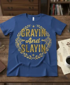 Grayin And Slayin quote with gold distressed font and laurel wreath with lion heads on a royal blue t-shirt