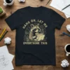 T-shirt design with text "Hang On, Let Me Overthink This" featuring a raccoon wearing glasses using a laptop and holding