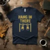Hang In There quote with golden gauntlets gripping a pull-up bar on a navy t-shirt