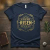 Navy blue t-shirt with gold text "HE IS NOT HERE RISEN JUST AS HE SAID" in a hexagonal design, featuring