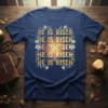 Navy blue t-shirt with golden "HE IS RISEN" text and a glowing cross graphic with floral accents