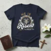 Navy blue t-shirt with "HE IS Risen matthew 28:6" text and a cross surrounded by a crown of thorns graphic