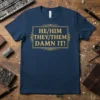 HE/HIM THEY/THEM DAMN IT! text in a golden rectangle on a navy blue t-shirt, promoting pronoun visibility