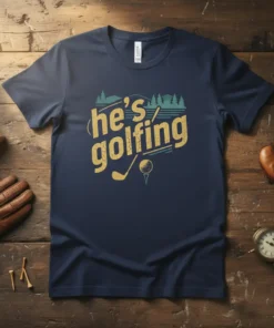 He's golfing t-shirt with distressed yellow text, golf club, and ball graphic, set against a backdrop of trees and water