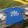 Blue t-shirt with white text "HI HUNGRY, I'M DAD" and graphics of a hot dog, burger, watermelon, and soda