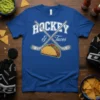 Blue t-shirt with white text "HOCKEY & Tacos" above crossed hockey sticks and a taco graphic