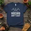 Hoeing Ain't Easy quote with crossed garden tools and a seedling graphic on a navy blue t-shirt