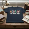 HOORAY! It's Library Day text design in colorful block letters with small star accents on a navy blue t-shirt