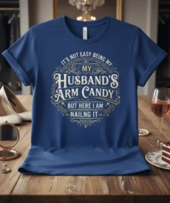 Blue t-shirt with gold and white text reading "It's Not Easy Being My Husband's Arm Candy But Here I Am