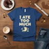 I Ate Too Much quote on a blue t-shirt with a cartoon stomach wearing a party hat and holding food