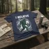 Navy blue t-shirt with white text "I BELIEVE" above a green Bigfoot graphic and forest design