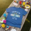 Blue t-shirt with "I Belong At The Kids Table" text and stick figures at a table graphic
