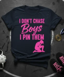 Pink text "I DON'T CHASE Boys I PIN THEM" with a female wrestler graphic on a dark navy t-shirt
