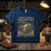 I Have Lived A Thousand Lives quote with an open book, forest, and city graphic on a navy blue t-shirt