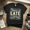Black t-shirt with white distressed text "I IDENTIFY AS LATE MY PRONOUNS ARE ALMOST/THERE" and a clock graphic