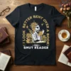 Black t-shirt with gold text: "I Look Better Bent Over A Book Proud Smut Reader", featuring a skeleton reading a