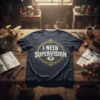 Dark navy t-shirt with the text "I NEED SUPERVISION" in distressed white font, surrounded by a decorative gold emblem and