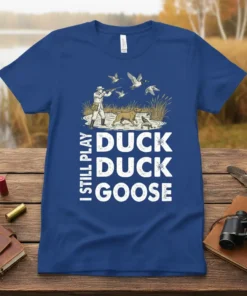 I Still Play Duck Duck Goose text design with hunter, dog, and flying ducks graphic, on a blue t-shirt