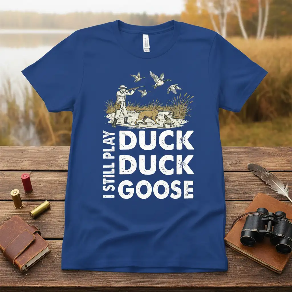 I Still Play Duck Duck Goose text design with hunter, dog, and flying ducks graphic on a blue t-shirt