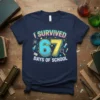 I SURVIVED 67 DAYS OF SCHOOL text with colorful numbers and school-themed graphics on a navy t-shirt