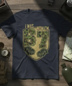 I WAS NOW I'M 67 military camouflage design on a navy blue t-shirt, featuring shield and hands graphic