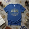 Blue t-shirt with gold text "I'M BUSY manifesting" and celestial graphics, ideal for manifestation and law of attraction enthusiasts