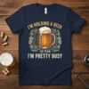 Navy blue t-shirt featuring the quote "I'm Holding A Beer - So Yeah - I'm Pretty Busy" with a graphic