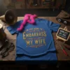 Blue t-shirt with gold glitter text that reads "I'm Just Here to Embarrass My Wife" in a decorative font