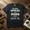 I'm Not Spoiled My Pops Just Loves Me quote in distressed white and gold text on a black t-shirt