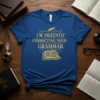 Blue t-shirt with gold text "I'M SILENTLY CORRECTING YOUR GRAMMAR." featuring a quill and an open book with a magnifying